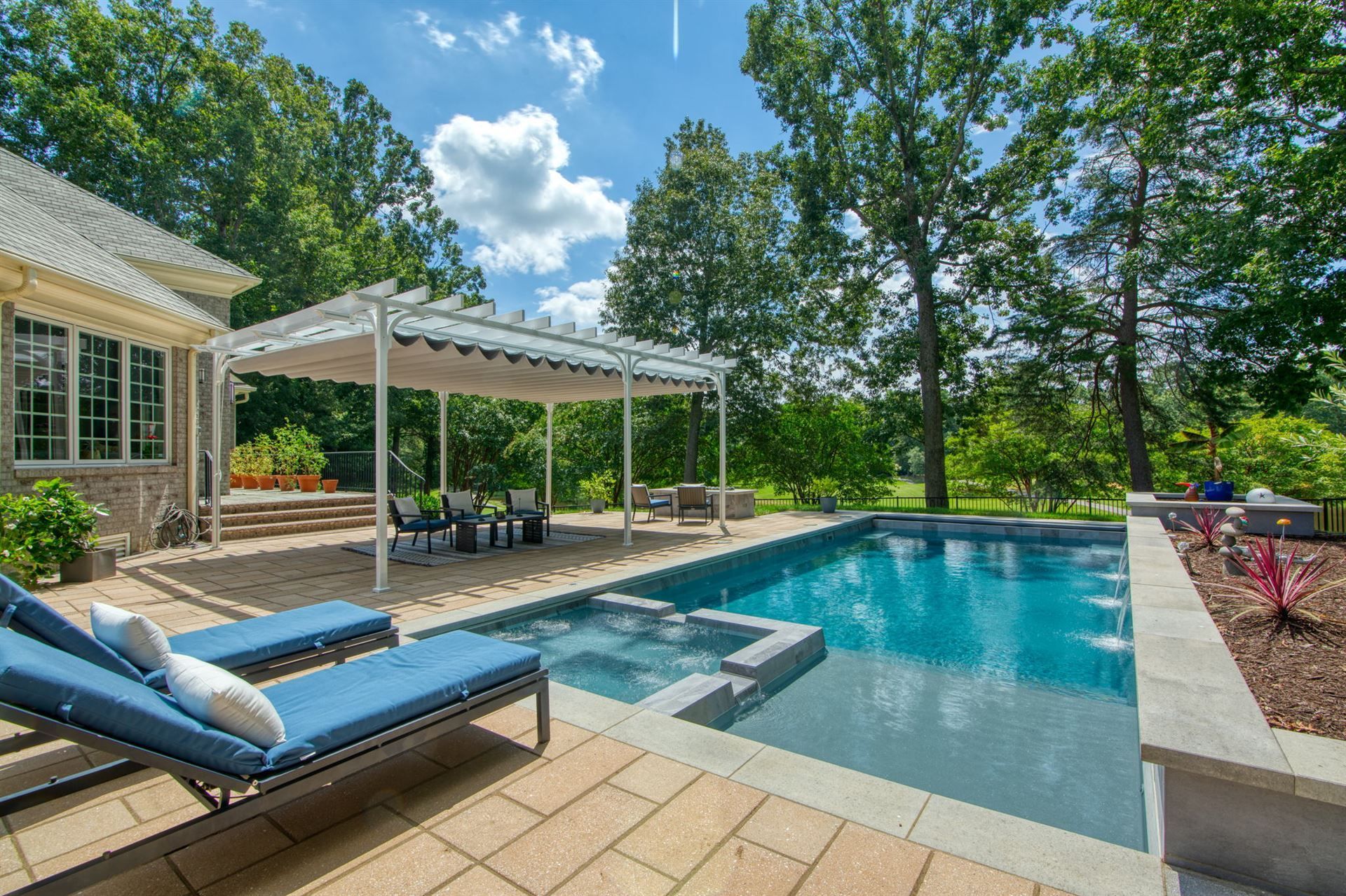 Backyard pool with spa, lounge chairs, and covered patio beside a stone house and trees