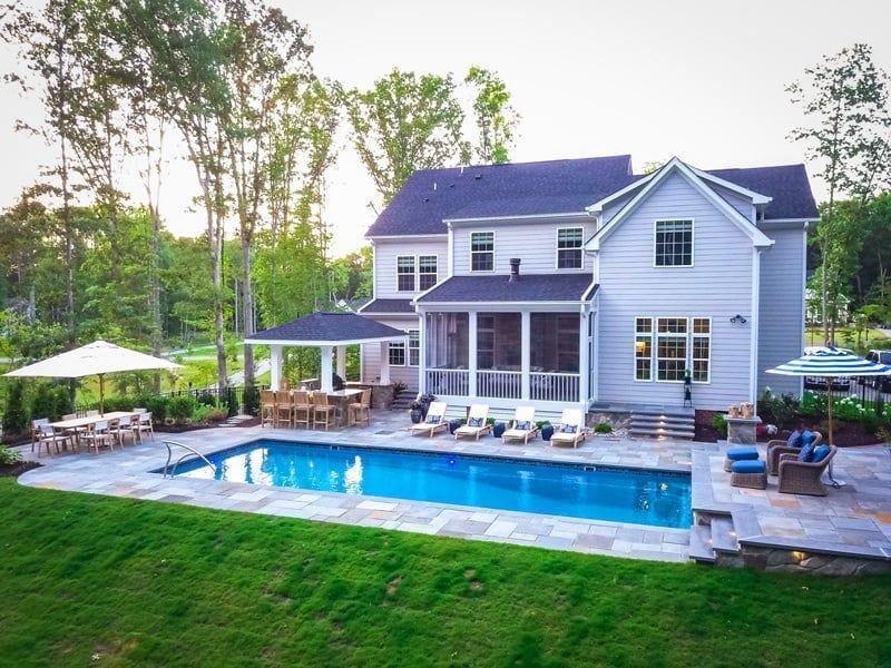 Backyard with modern white house, blue swimming pool, lounge chairs, and patio under trees