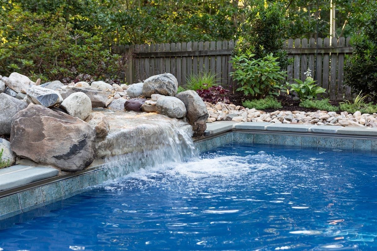 Backyard pool with a small rock waterfall and blue water, bordered by landscaping and a wooden fence