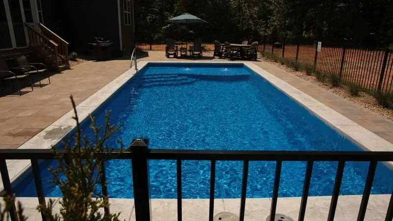 Rectangular outdoor swimming pool with blue water, surrounded by a stone deck and black fence in sunlight