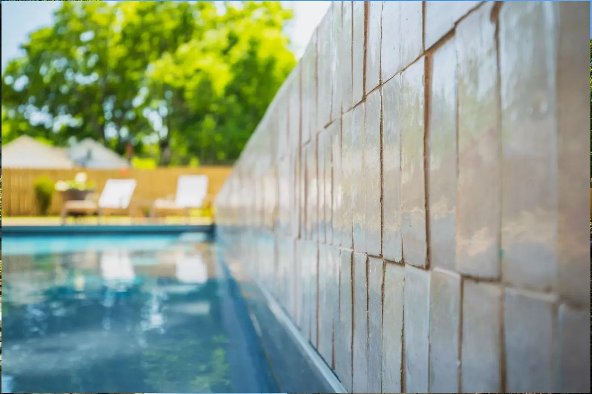 Poolside view with blue water beside a tiled wall and sunlit trees in the background