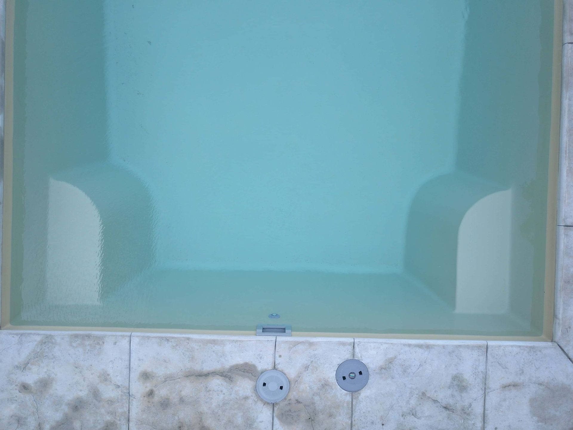 Blue-tinted bathtub with two faucets and white side panels behind frosted glass.