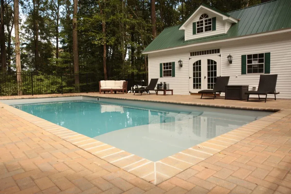 Backyard pool beside a white house with a green roof and brick patio in a wooded setting