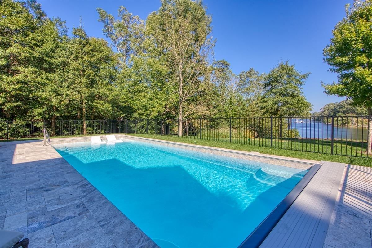 Rectangular backyard pool with turquoise water, stone deck, and trees by a black metal fence