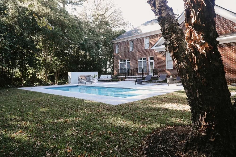 Backyard swimming pool beside a brick house, with lawn and trees in the foreground.