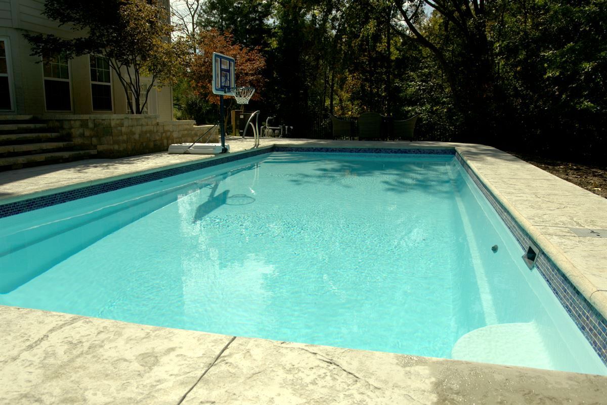Sunlit outdoor swimming pool with blue water and surrounding patio, trees in the background