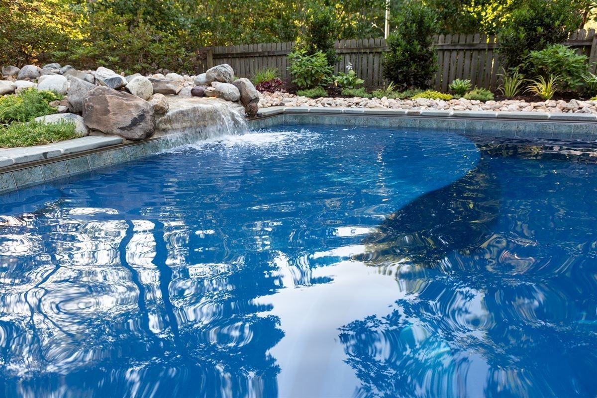 Backyard swimming pool with blue water, stone edge, and landscaped rocks and greenery.