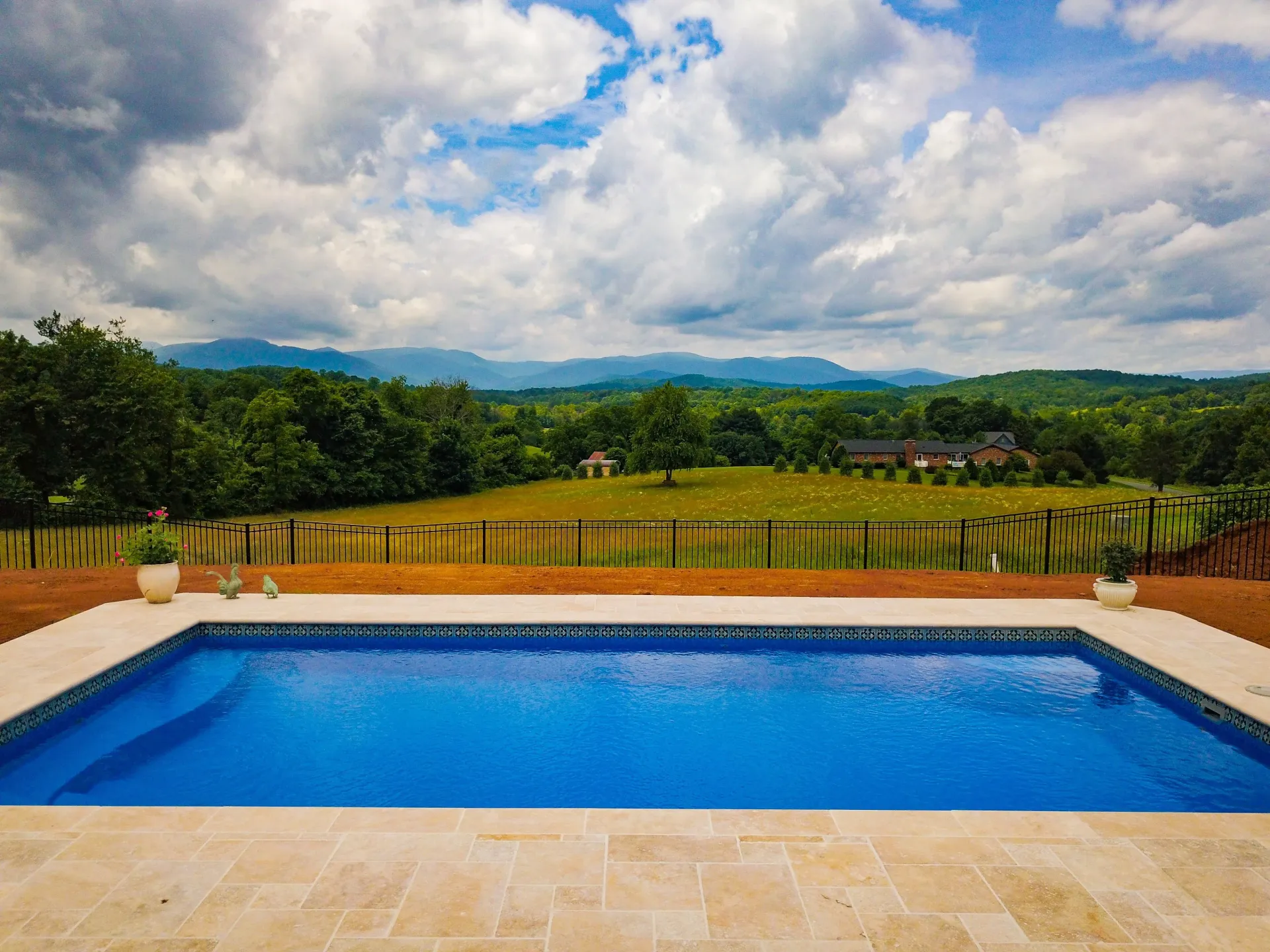 Backyard pool with blue water overlooking green fields and distant hills under a cloudy sky