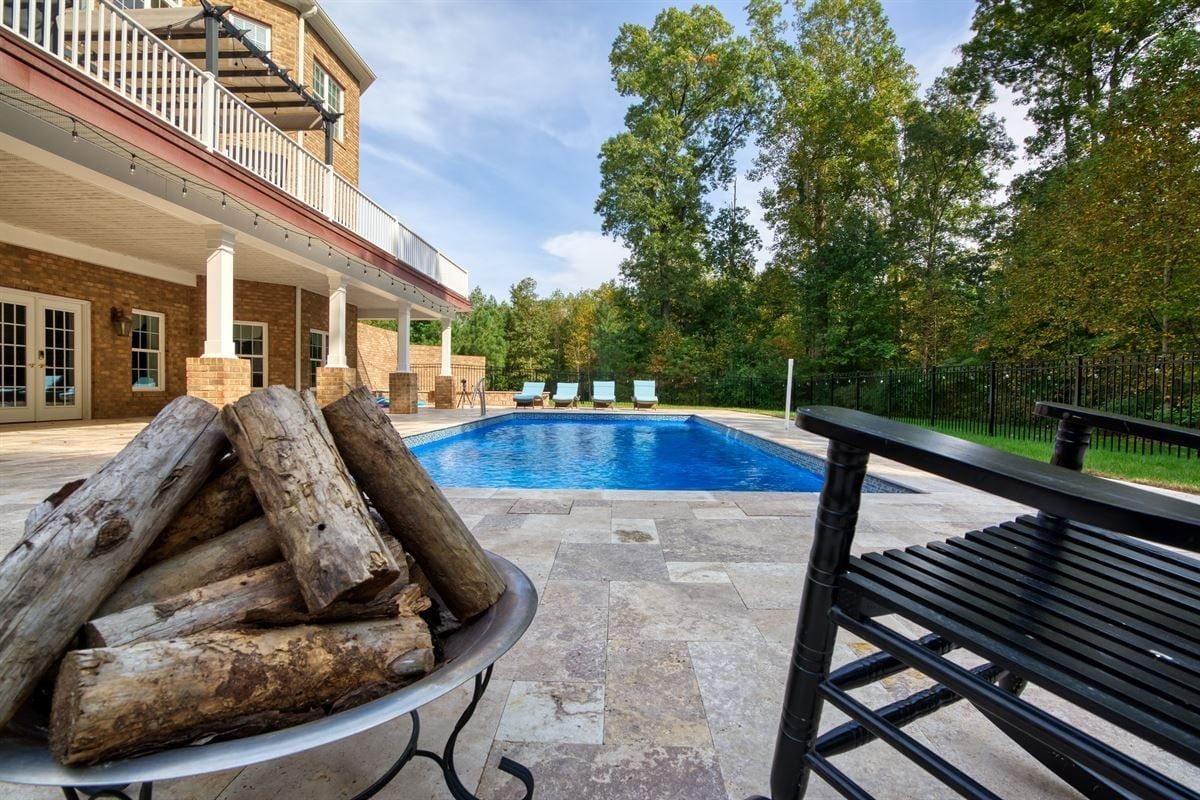 Backyard patio with a blue swimming pool, stacked firewood, and trees beyond a black metal fence