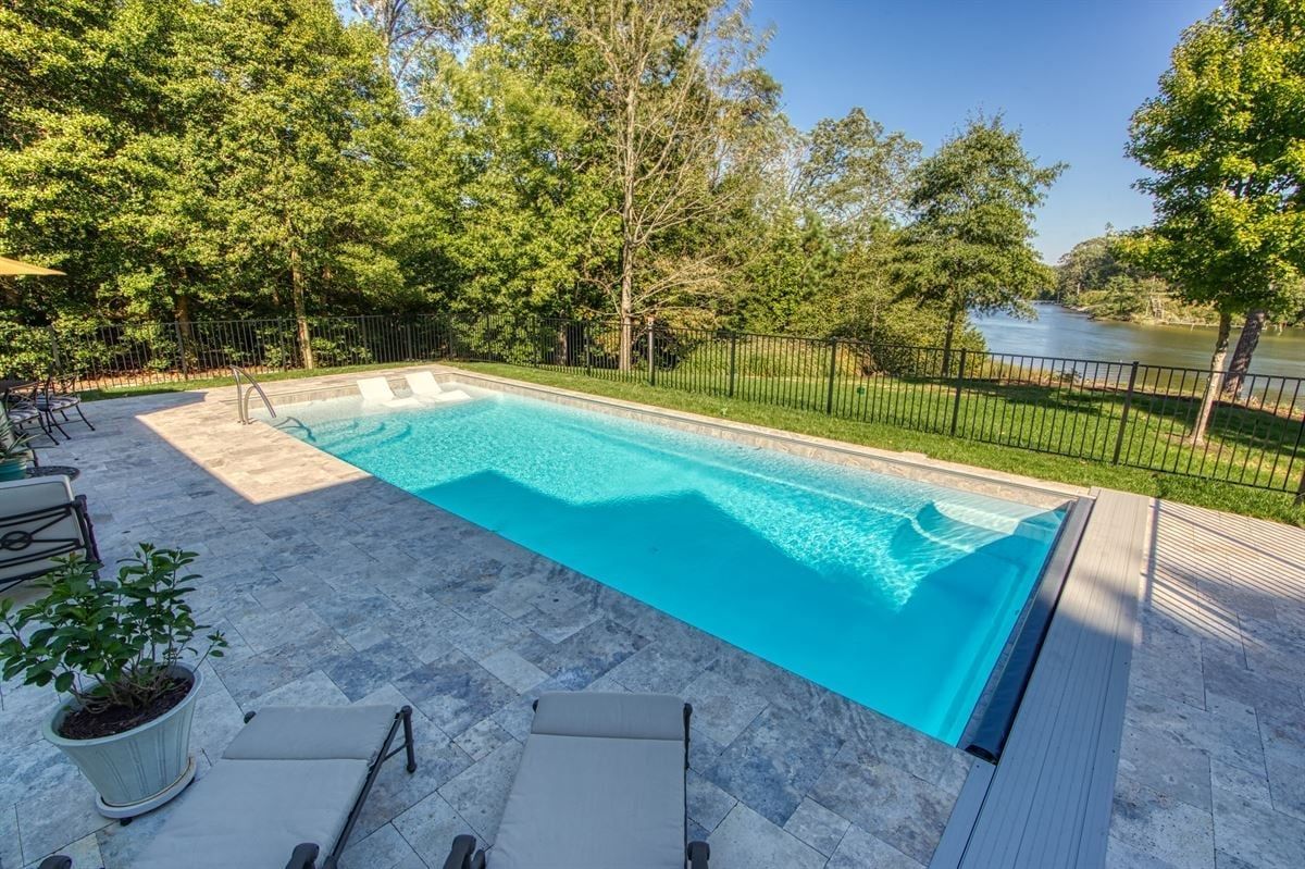 Backyard swimming pool with blue water, stone deck, and trees overlooking a lake