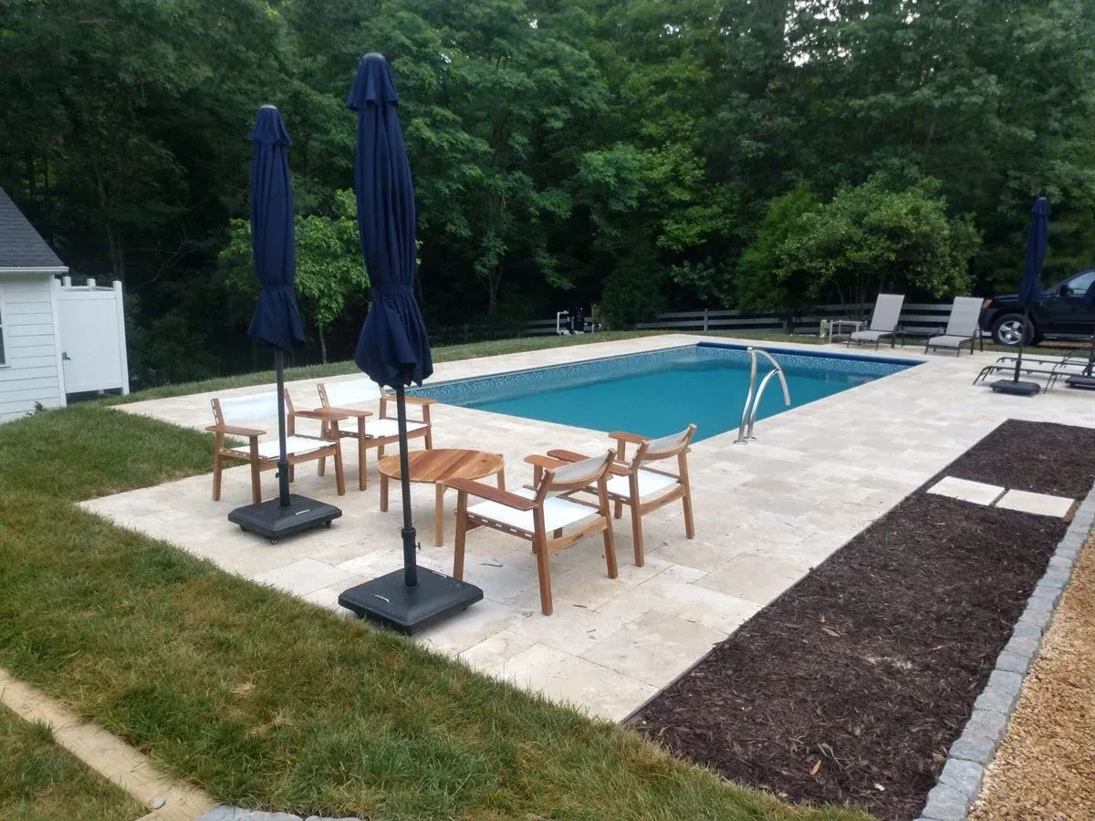 Backyard pool deck with wooden table and chairs, blue umbrellas, and lounge chairs beside a swimming pool.