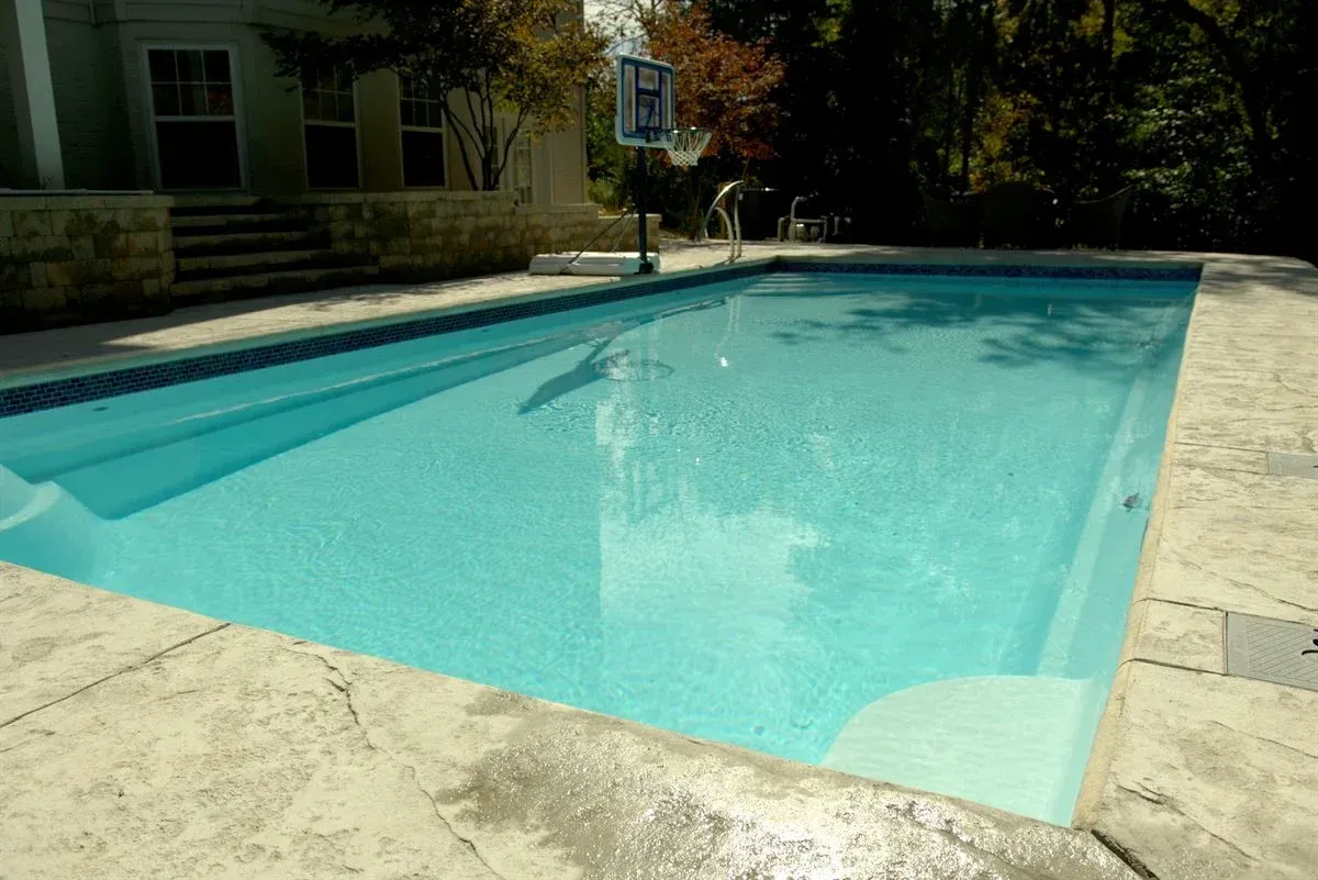 Turquoise swimming pool beside a stone patio, with a basketball hoop and house in the background