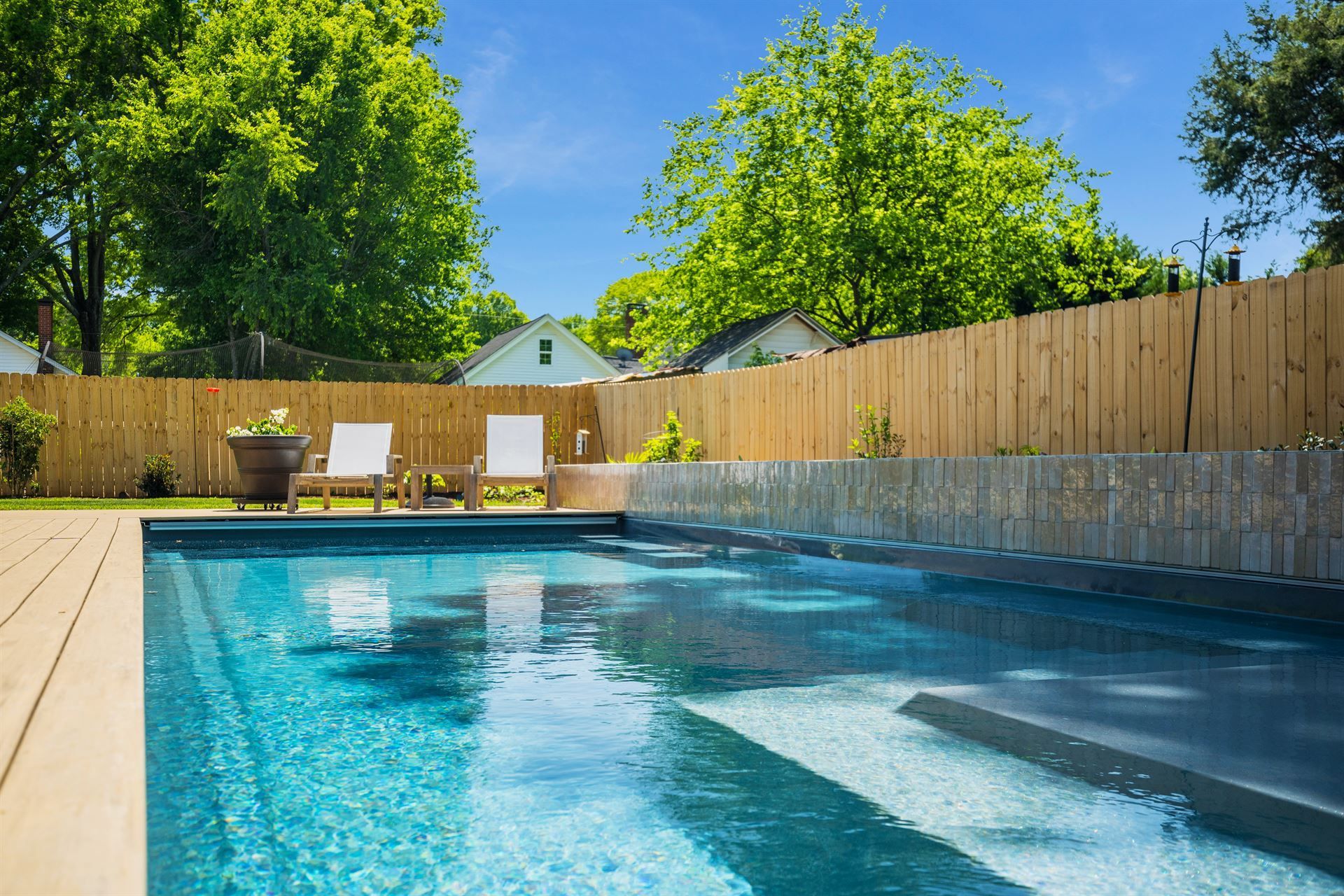 Backyard swimming pool with blue water, lounge chairs, wooden fence, and green trees under a bright sky