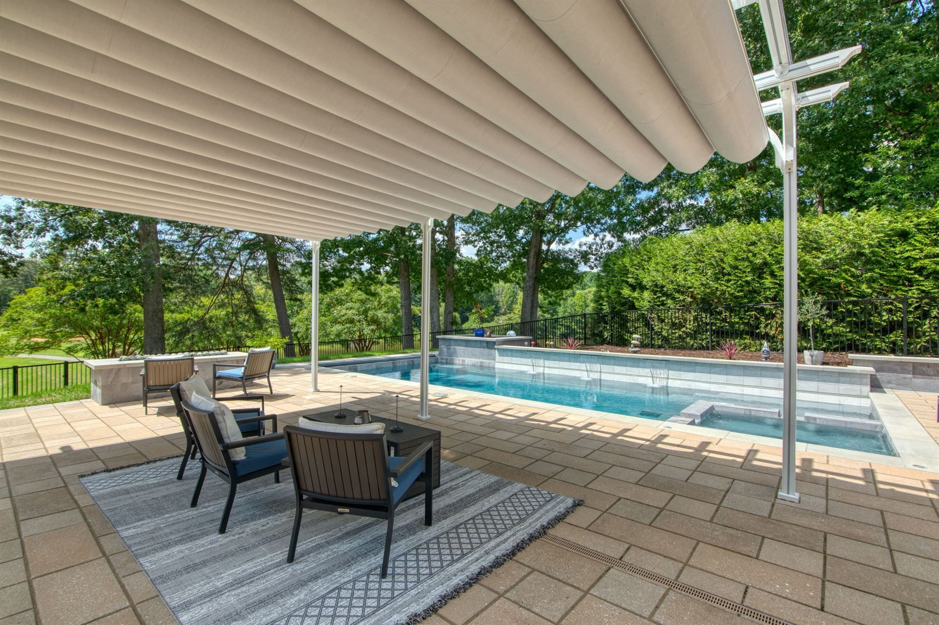 Covered patio with lounge chairs beside a backyard swimming pool and trees beyond