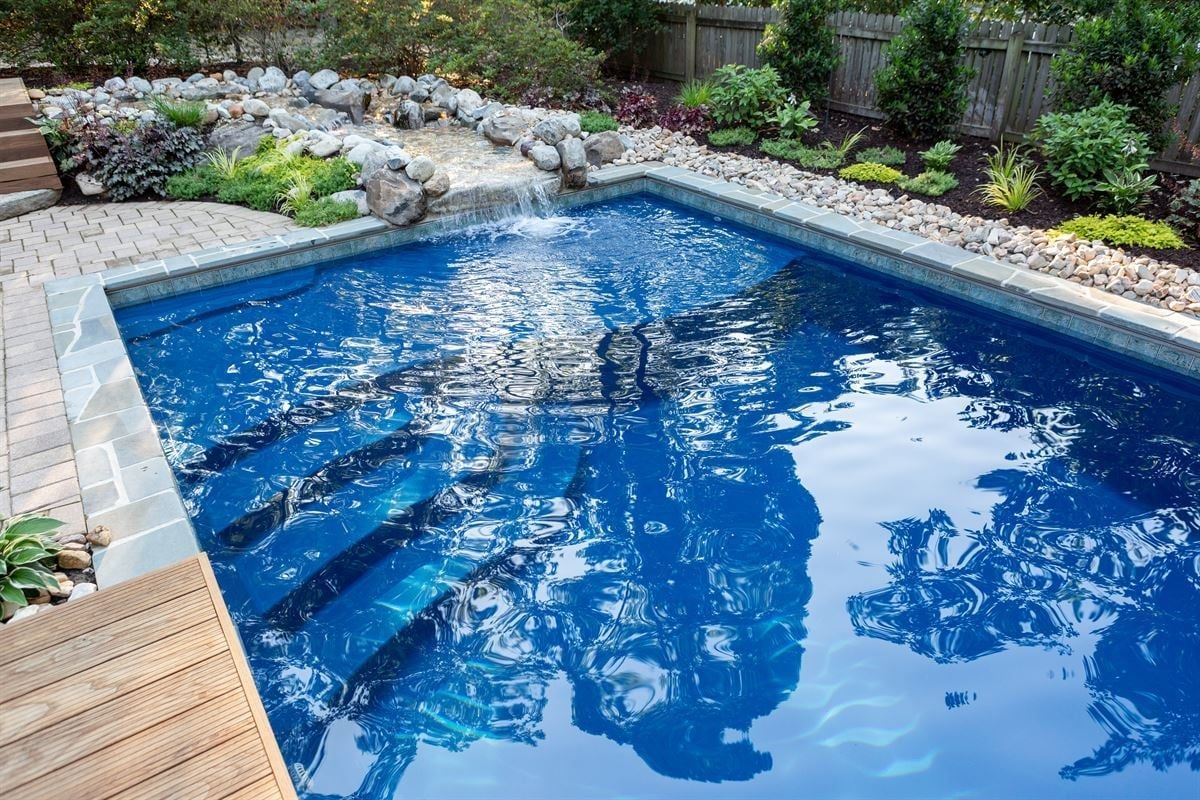 Blue backyard swimming pool with stone edging, wooden deck, and landscaped garden border