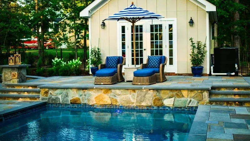 Backyard pool beside a cream cabana with blue lounge chairs and striped umbrella