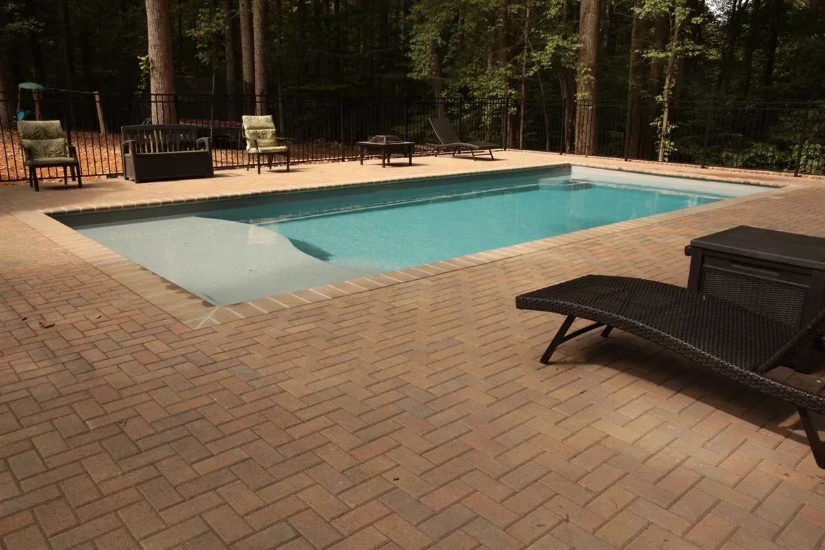 Brick patio with a rectangular swimming pool and lounge chairs in a wooded backyard