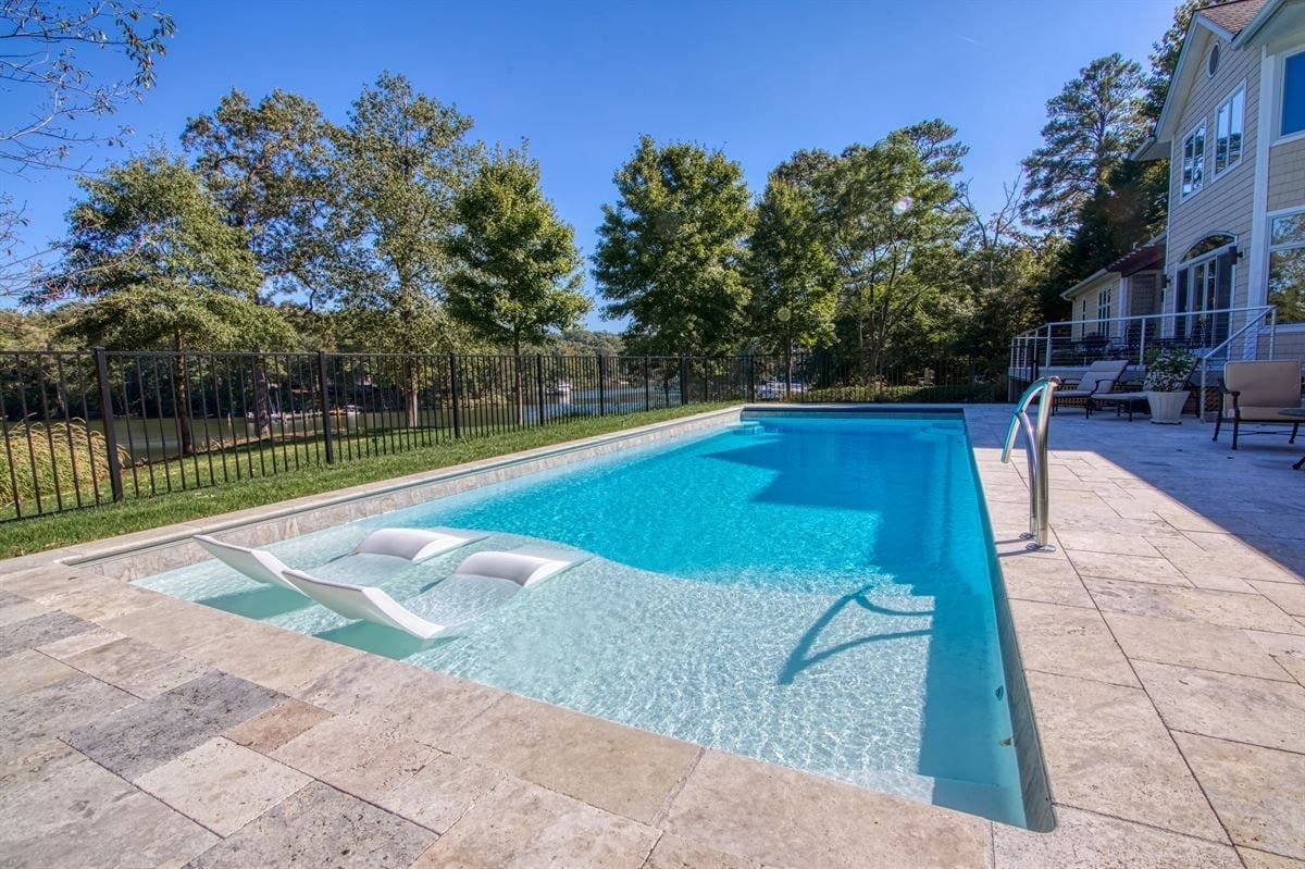 Outdoor swimming pool with clear blue water, stone deck, lounge chairs, and trees under a sunny sky