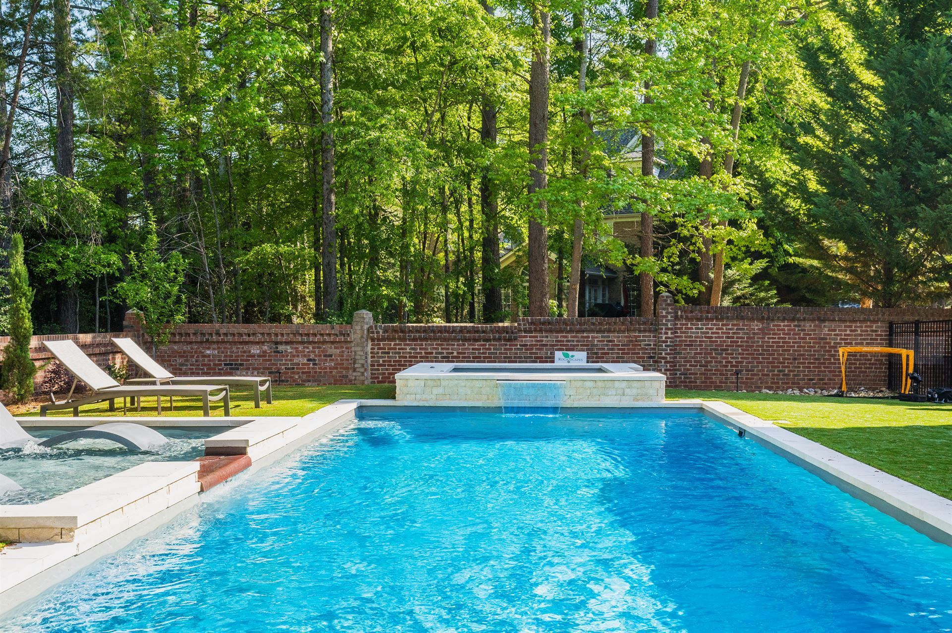 Outdoor pool with blue water, lounge chairs, and a stone wall backed by tall green trees