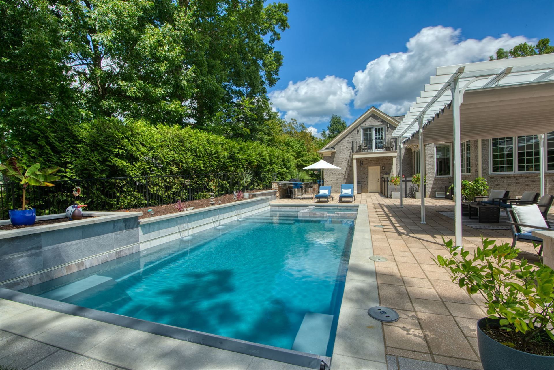 Backyard pool beside a house with patio chairs and lush green trees under a blue sky
