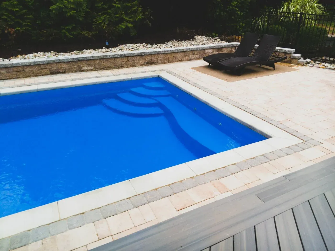 Blue swimming pool beside a stone deck with a black lounge chair and greenery beyond