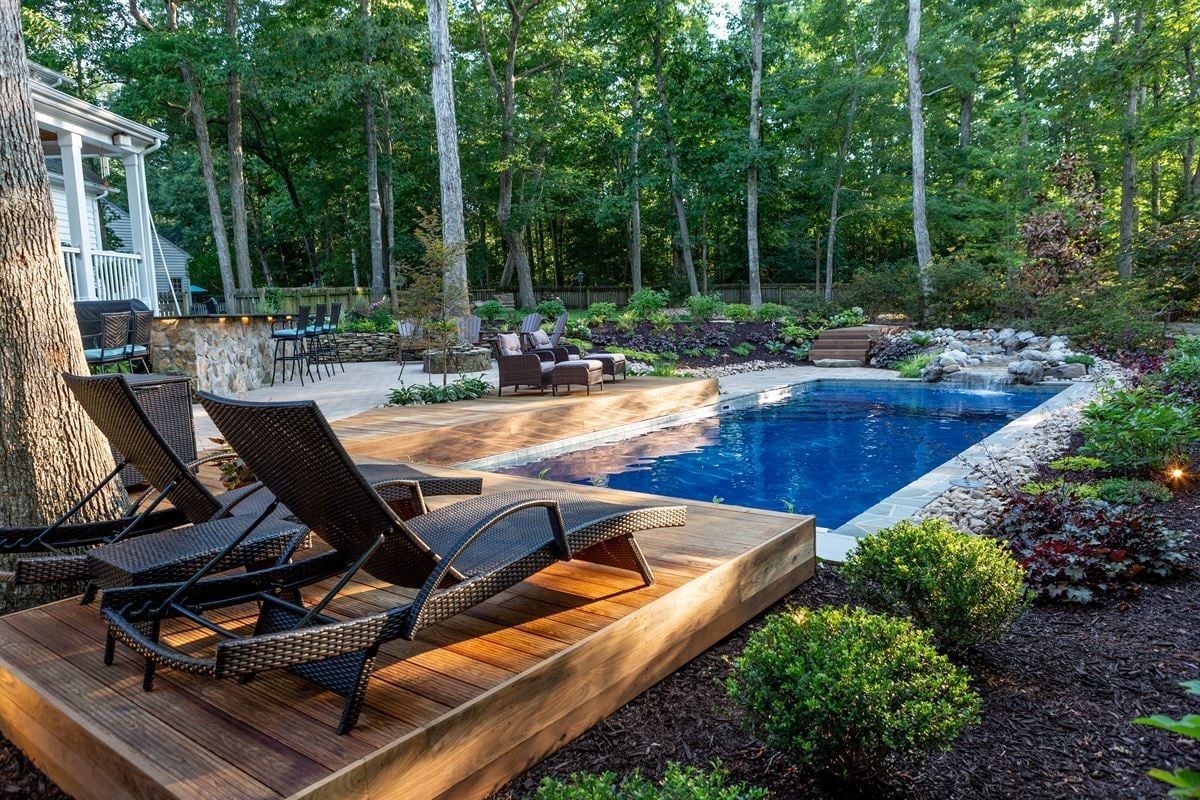 Backyard pool with wooden deck chairs, stone patio, and lush green landscaping