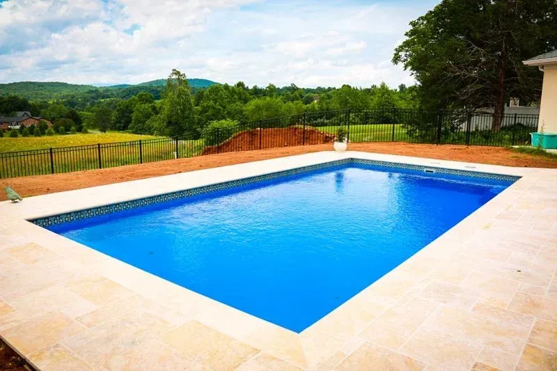 Rectangular blue swimming pool beside a tan patio with green trees and hills in the background
