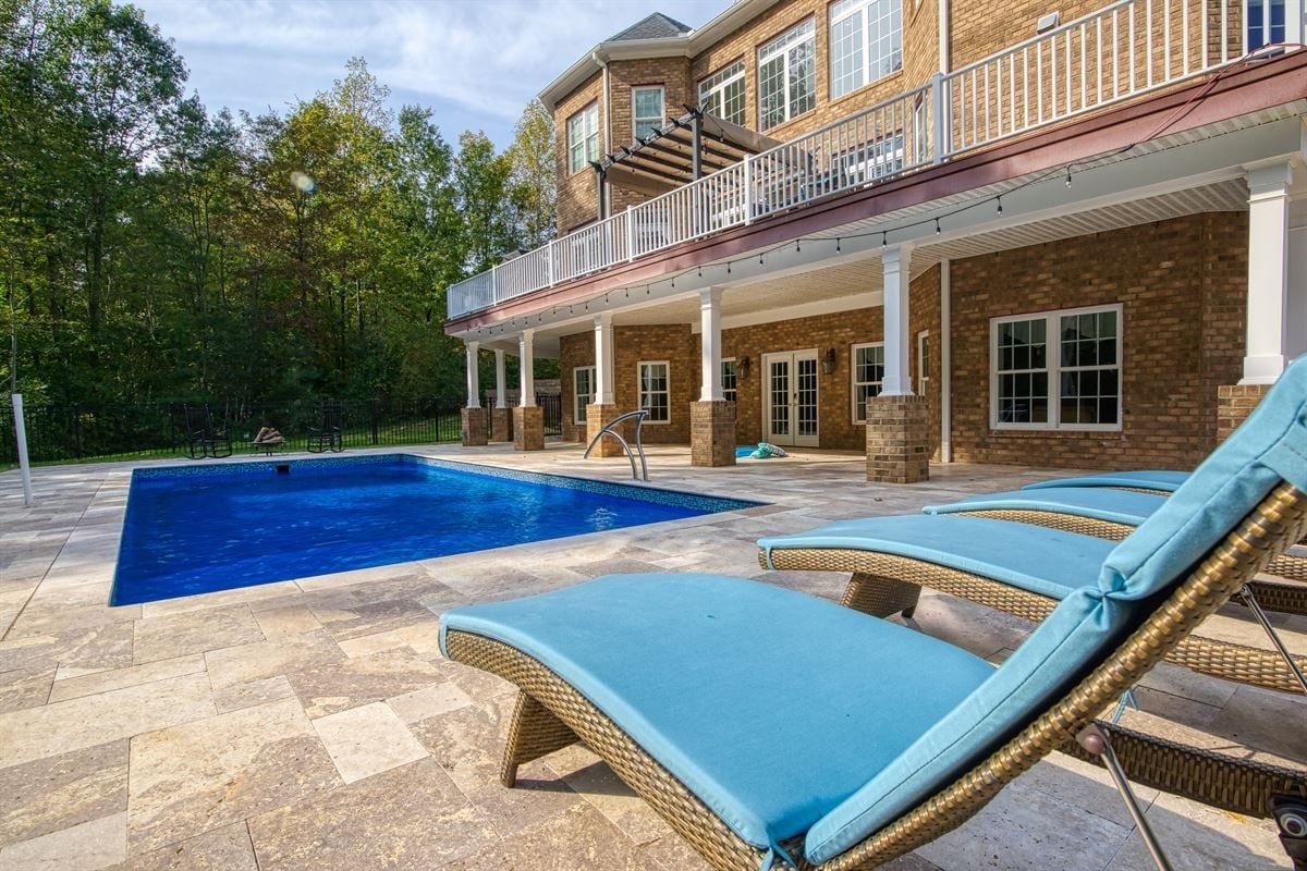 Backyard patio with blue pool and cushioned lounge chairs beside a brick house