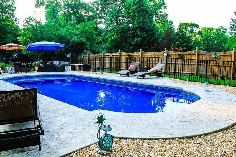 Backyard swimming pool with lounge chairs, blue umbrella, and fenced patio surrounded by trees