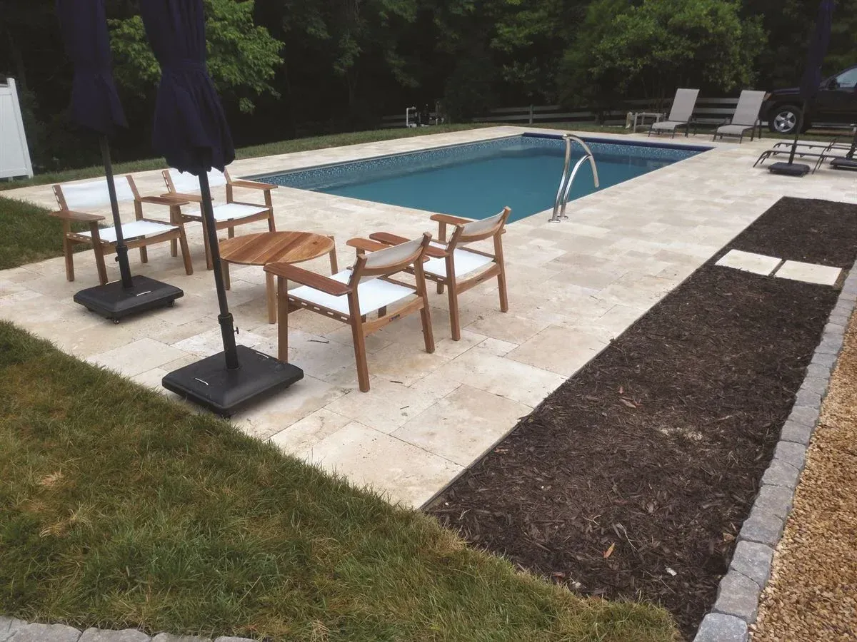 Patio seating beside a swimming pool, with wooden chairs and tables on a stone deck