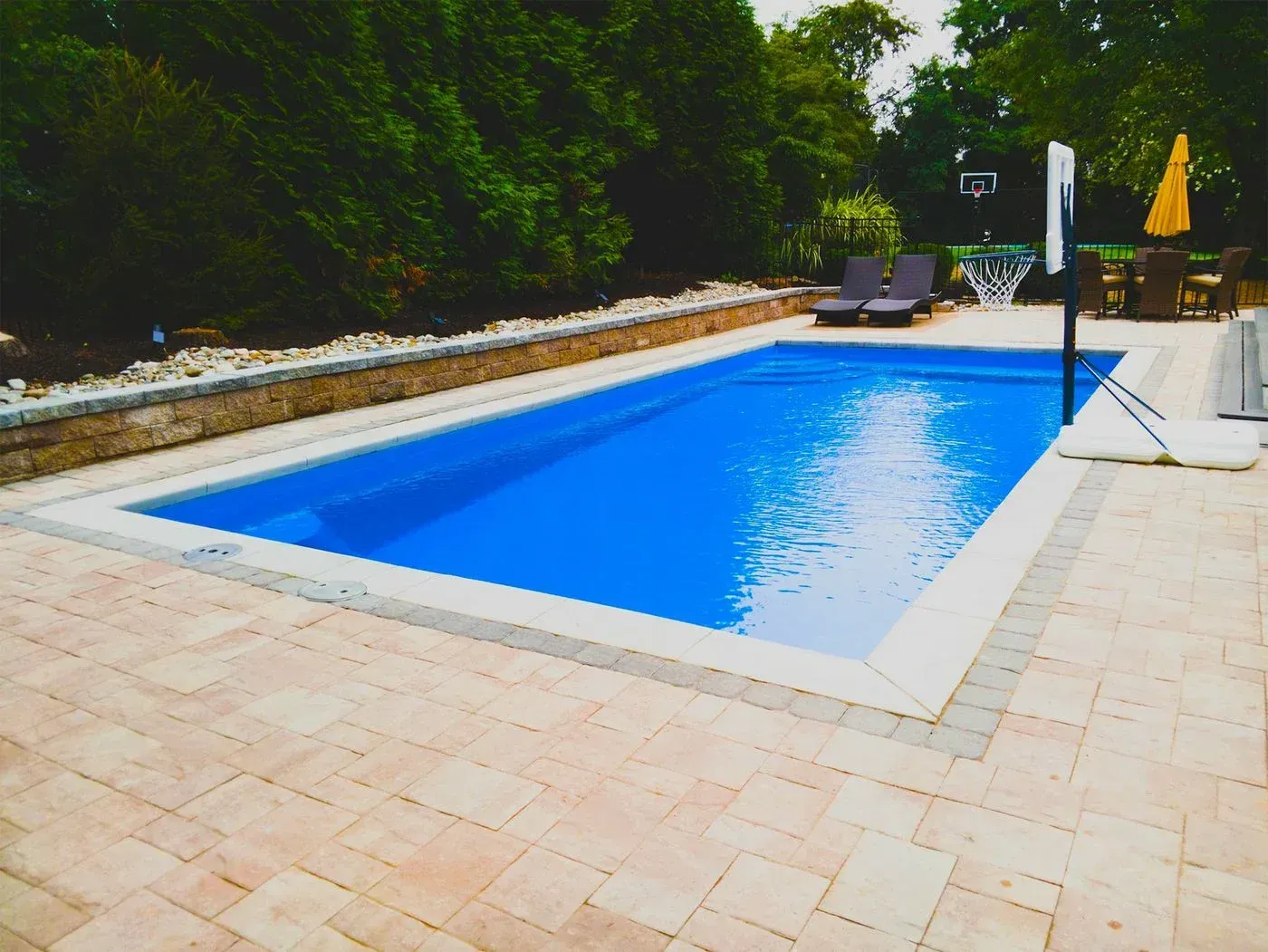 Backyard swimming pool with blue water, stone patio, lounge chairs, and green trees around it
