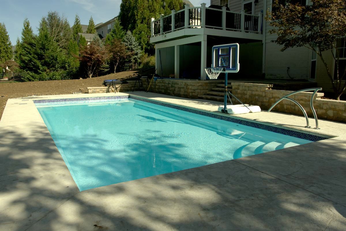 Backyard swimming pool beside a house, with a portable basketball hoop and trees nearby