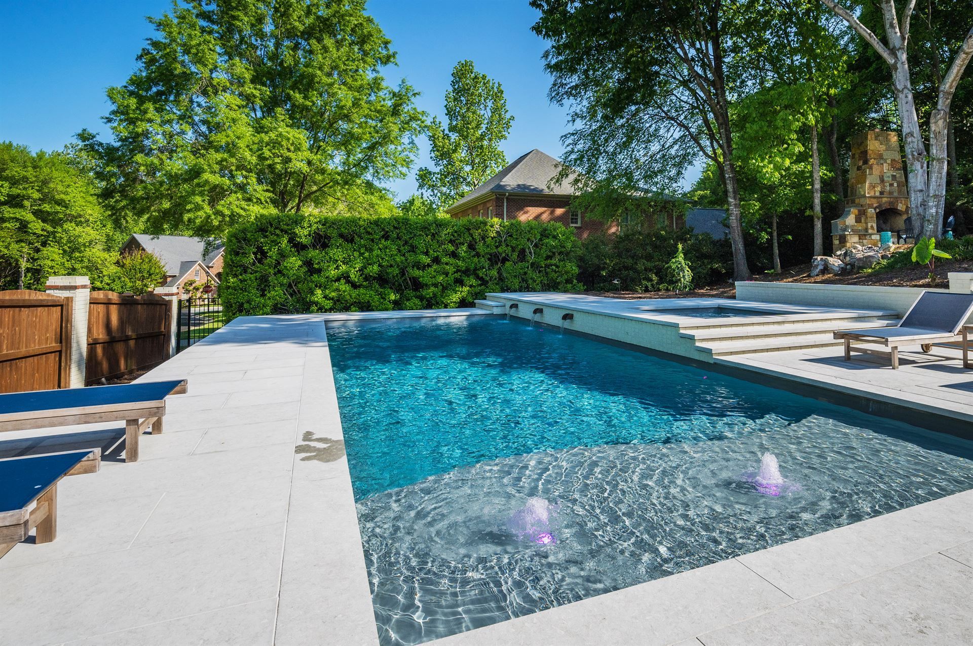 Backyard pool with blue water, white deck, lounge chairs, and lush green trees under a sunny sky