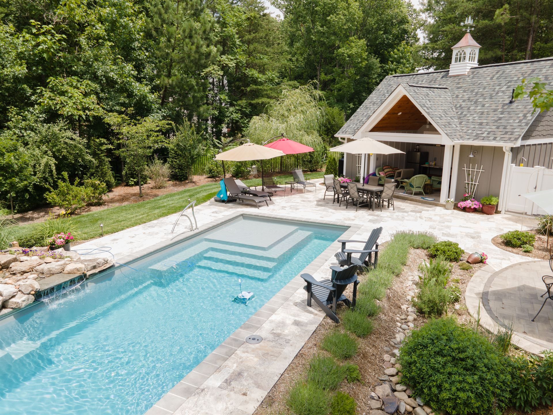Backyard pool with lounge chairs, umbrellas, and a stone cottage patio surrounded by greenery