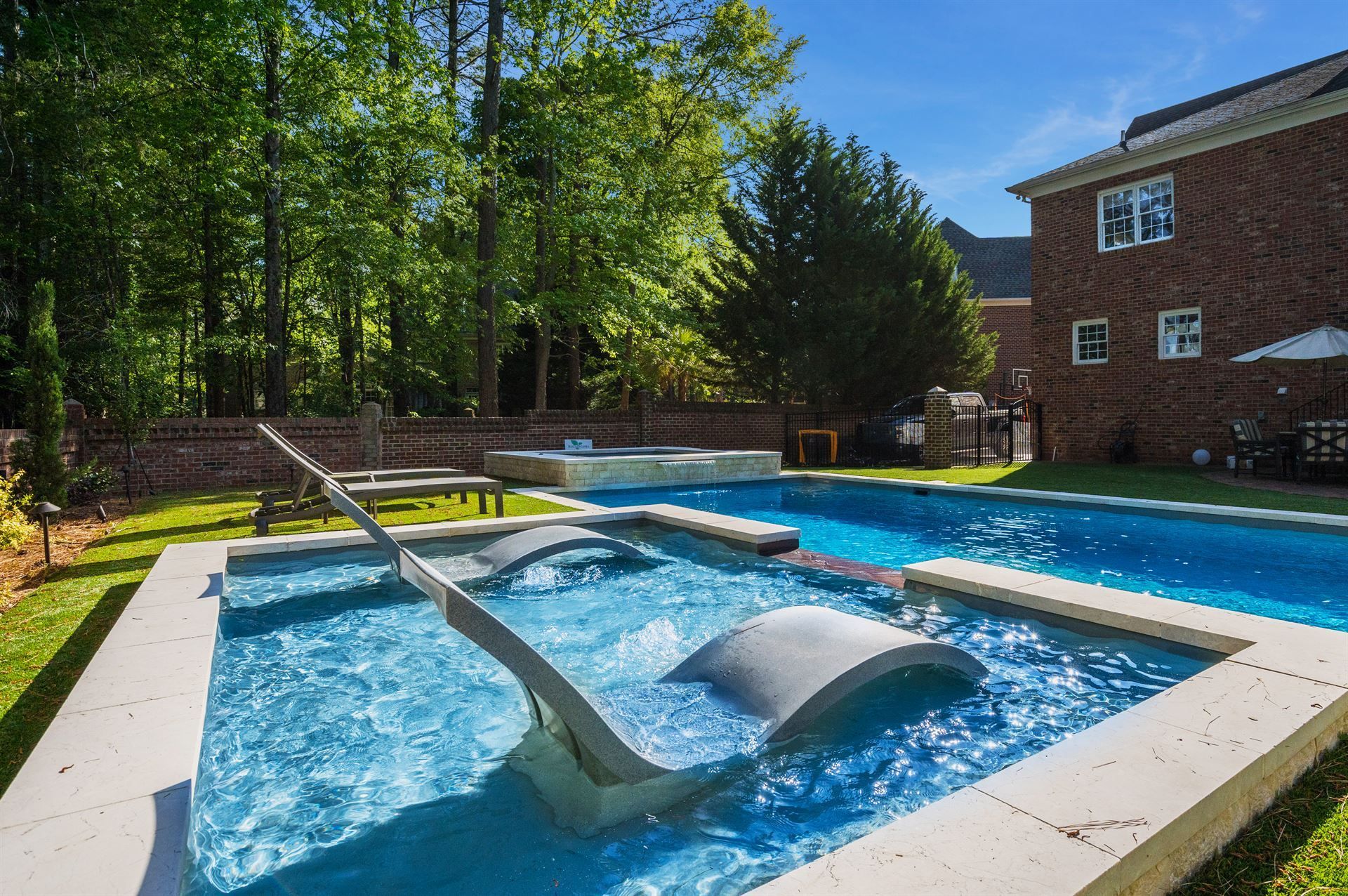Backyard pool with fountain jets beside a brick house and trees in bright sunlight