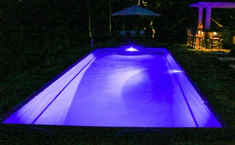 Glowing purple-lit swimming pool at night with umbrellas and a lit seating area in the background
