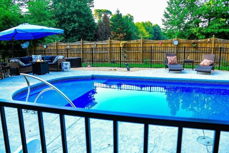 Backyard pool with blue water, patio chairs, umbrella, and wooden fence surrounded by greenery