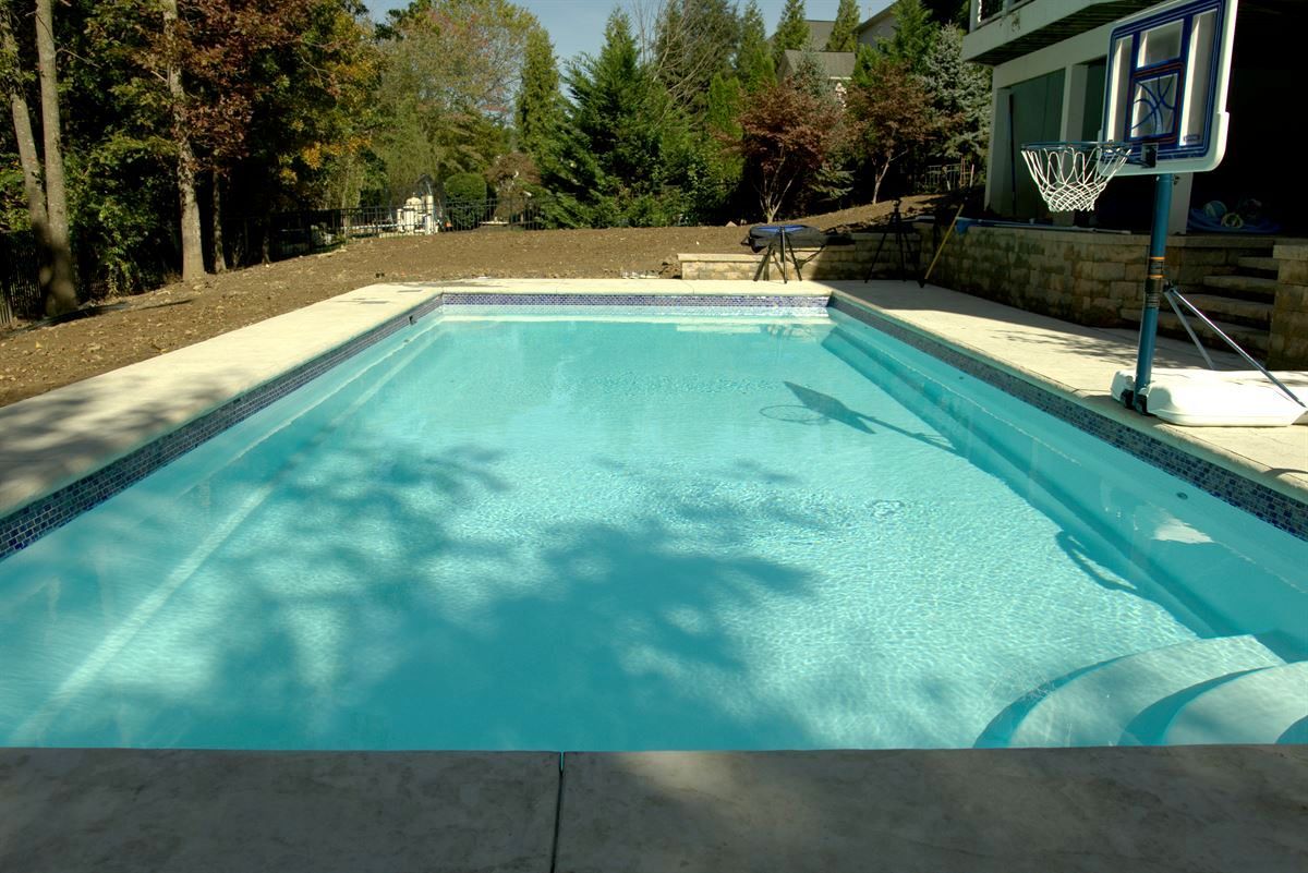 Backyard swimming pool with clear blue water, surrounded by concrete patio and trees.