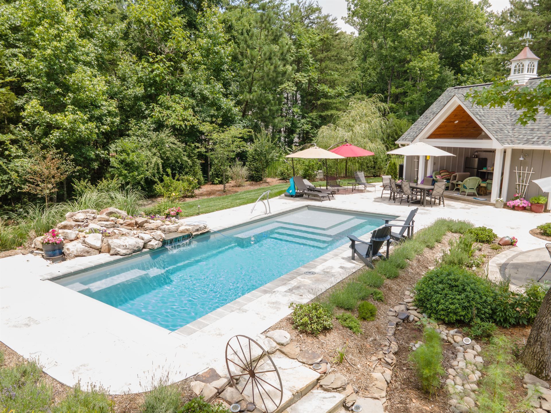 Backyard pool with lounge chairs, patio umbrellas, and a stone pool house surrounded by trees