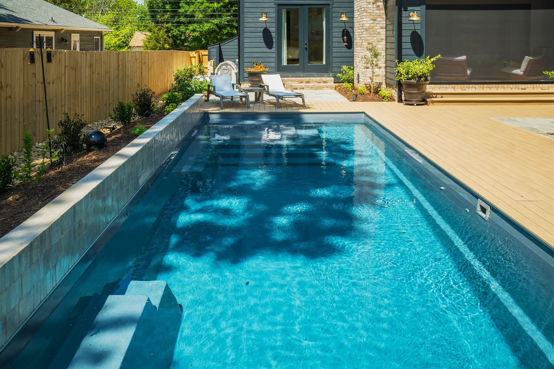 Long narrow backyard swimming pool with blue water, patio seating, and a modern house in the background