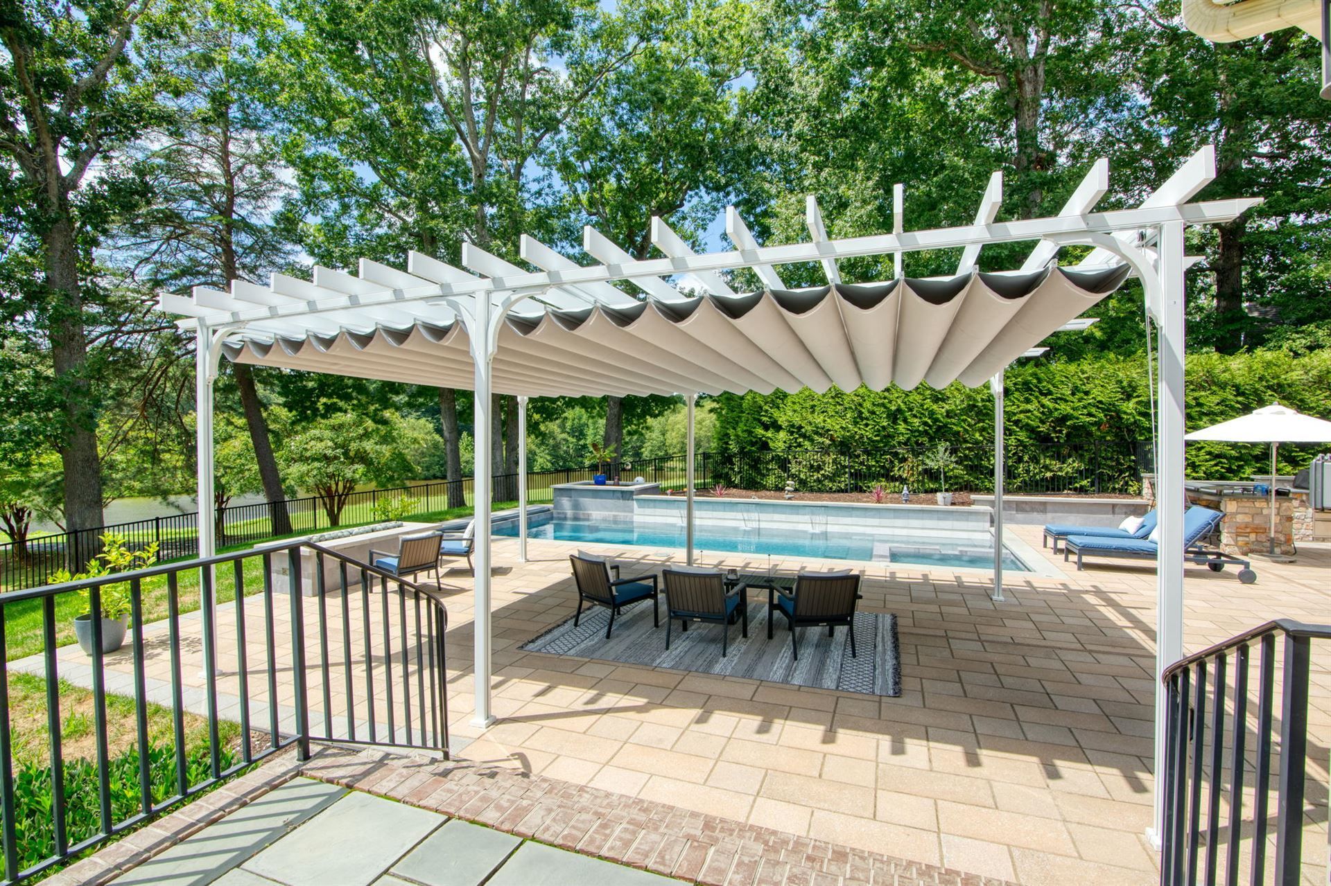 Patio under a white pergola beside a backyard pool, with lounge chairs and trees in the background