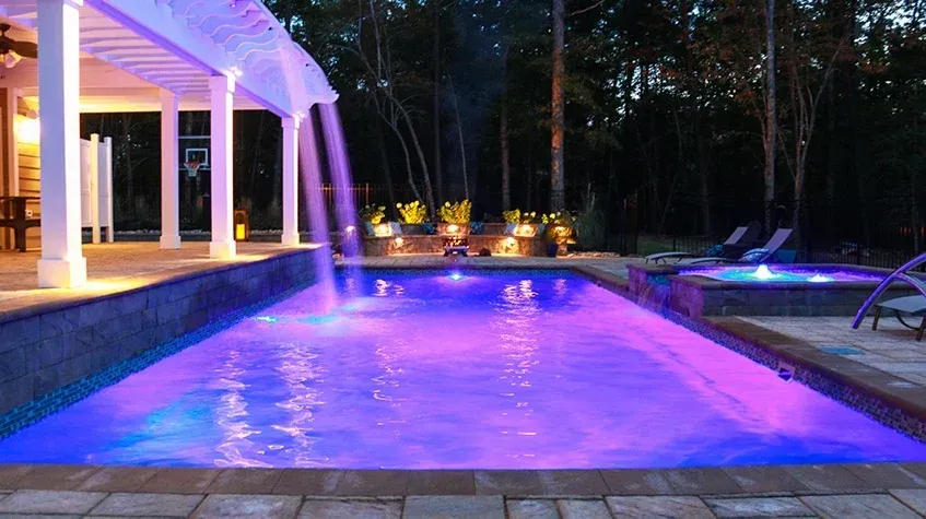 Purple-lit outdoor pool beside a modern patio at night, with trees and lounge chairs in the background