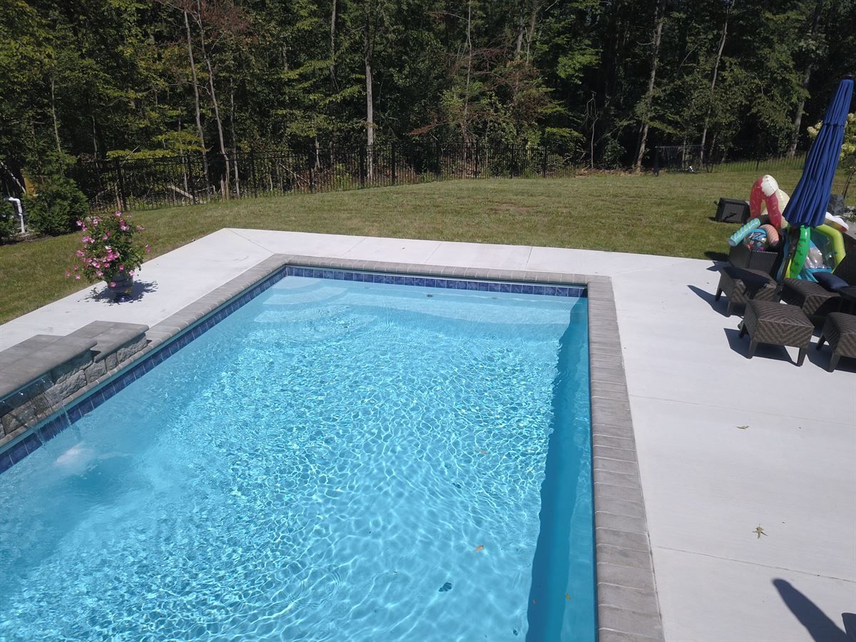 Backyard swimming pool with blue water, concrete deck, and lounge chairs by a wooded yard