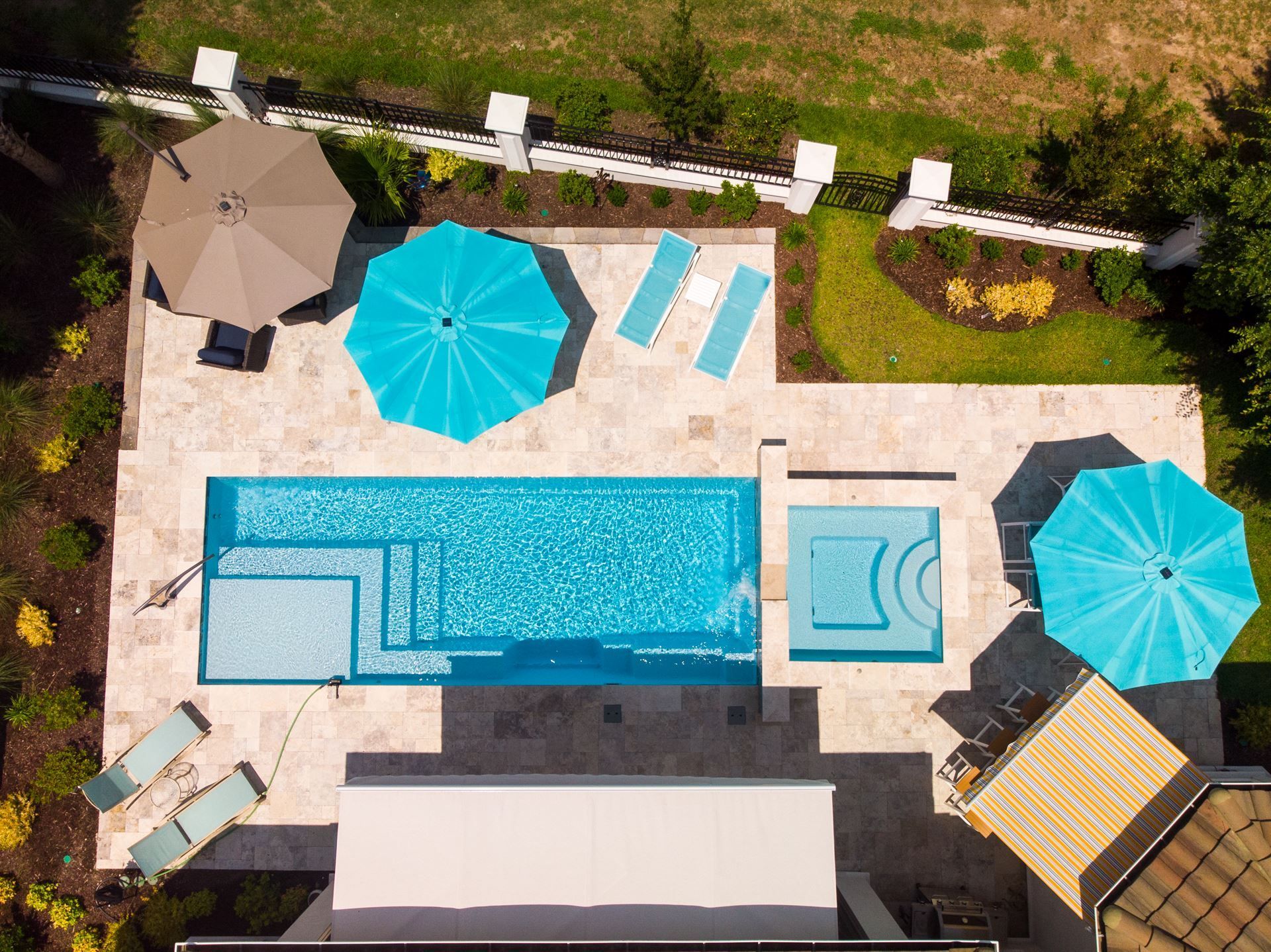 Aerial view of a backyard with a rectangular pool, hot tub, turquoise umbrellas, and lounge chairs.