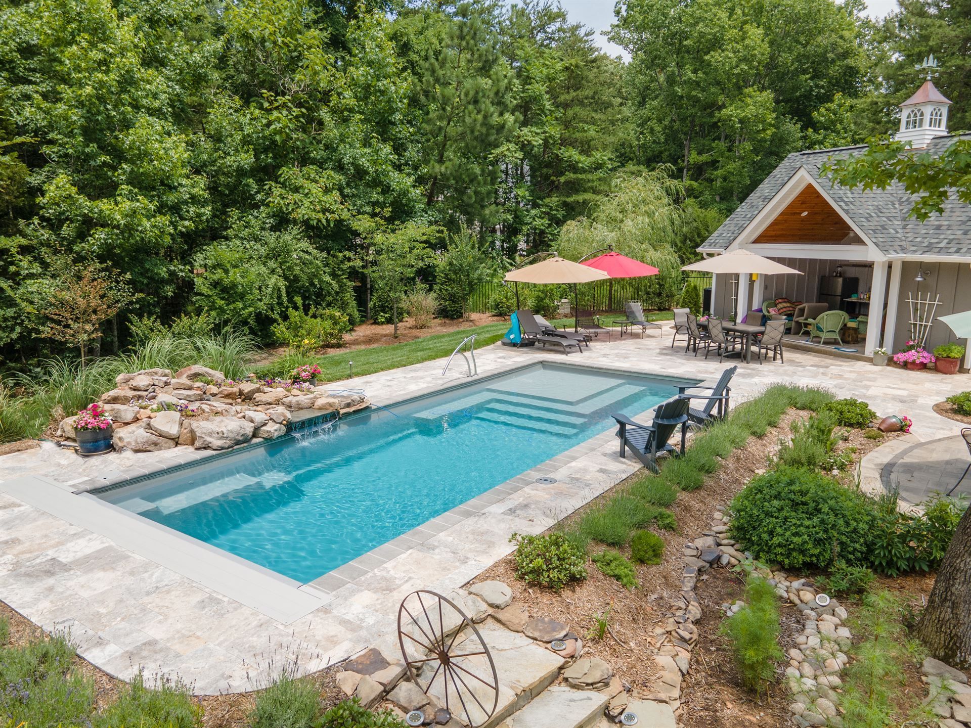 Backyard pool with lounge chairs, umbrellas, and a patio beside a small pool house, surrounded by greenery