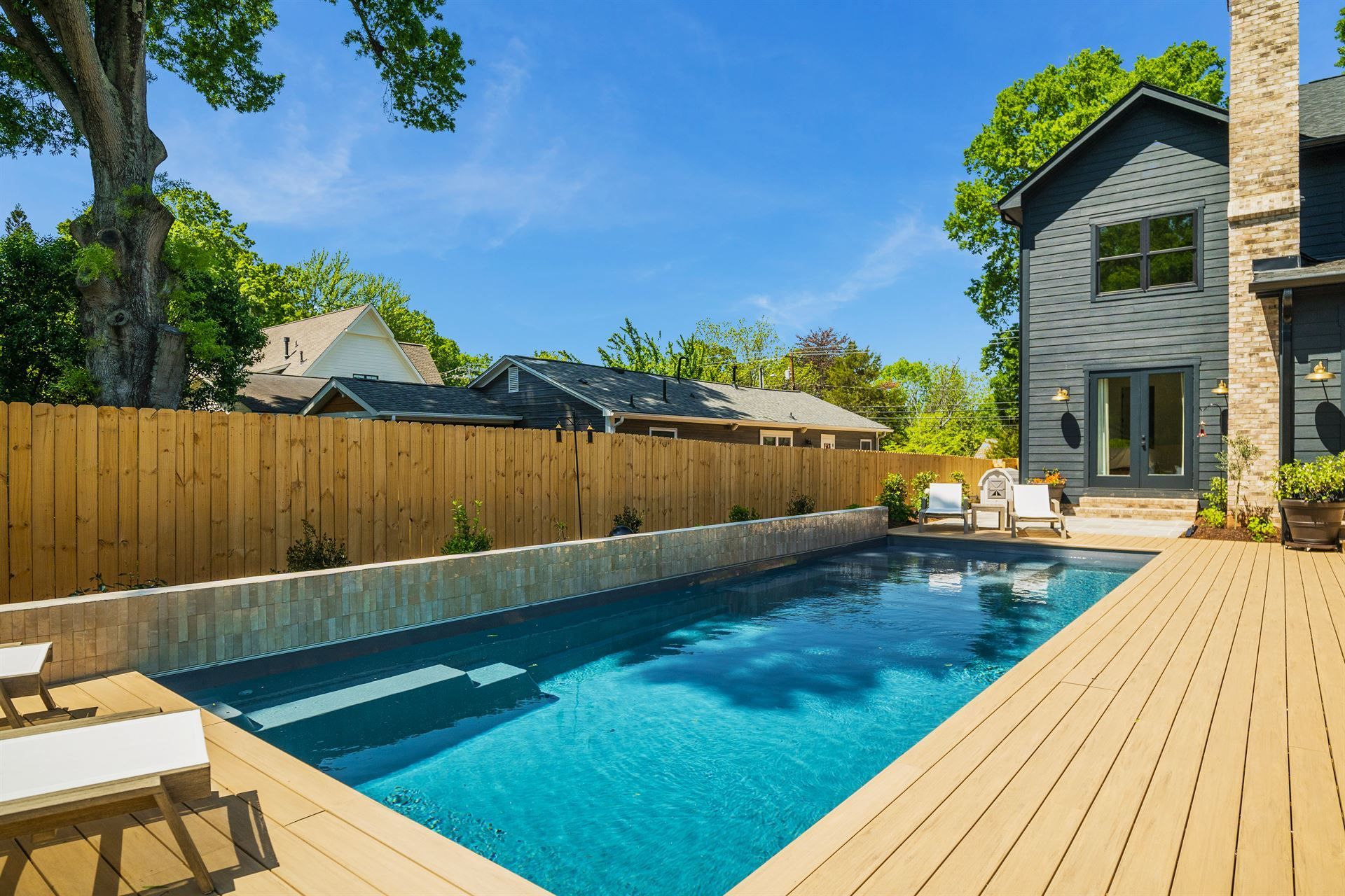 Backyard with a rectangular swimming pool, wooden deck, privacy fence, and house under a blue sky
