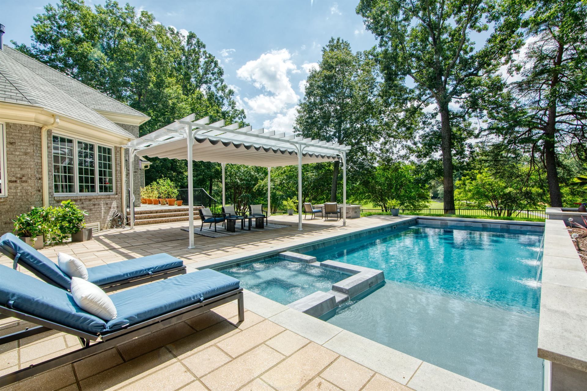 Backyard pool with lounge chairs, covered patio, and stone house surrounded by trees