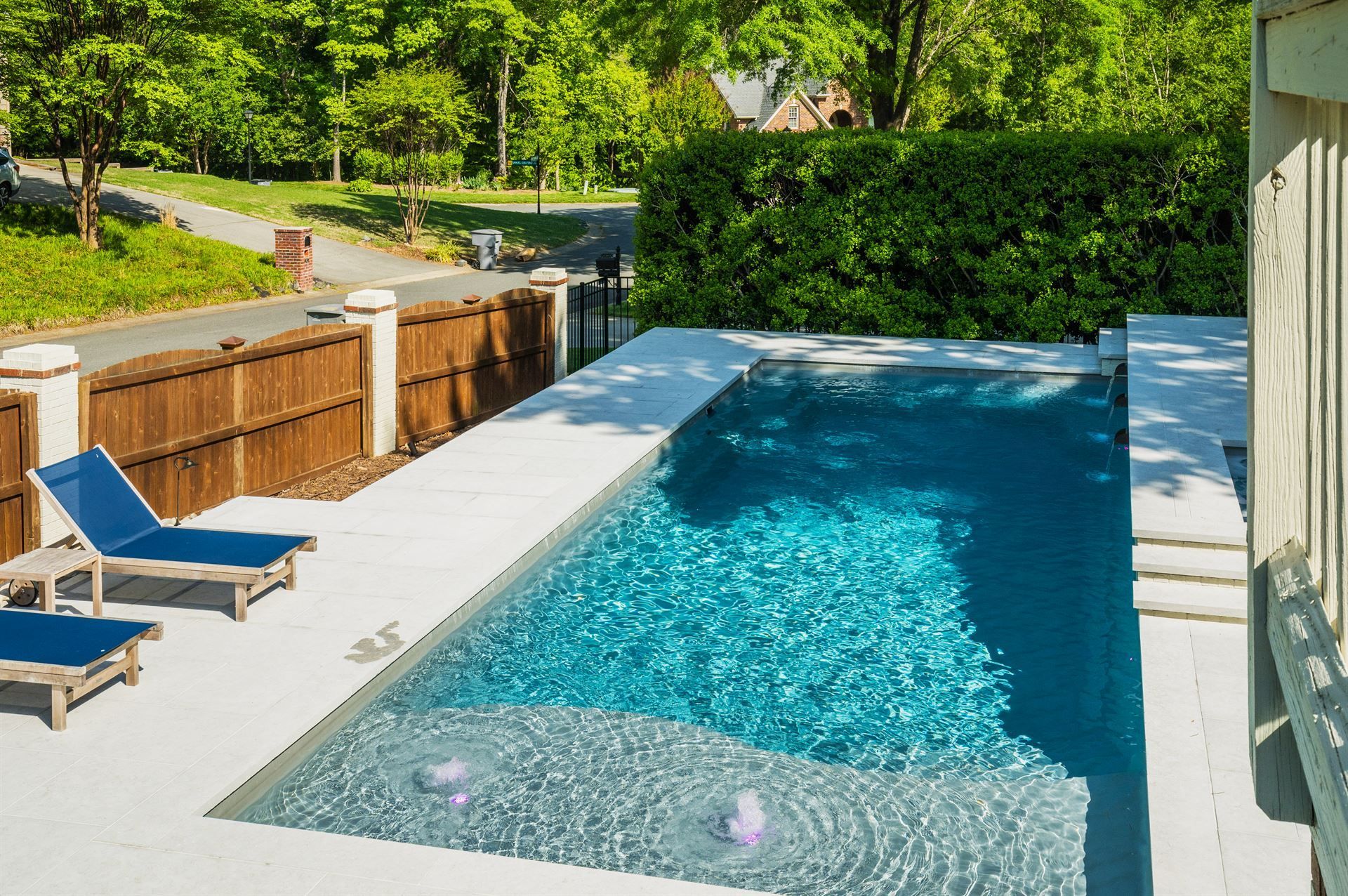 Rectangular backyard pool with blue water, lounge chairs, and a wooden fence in bright sunlight