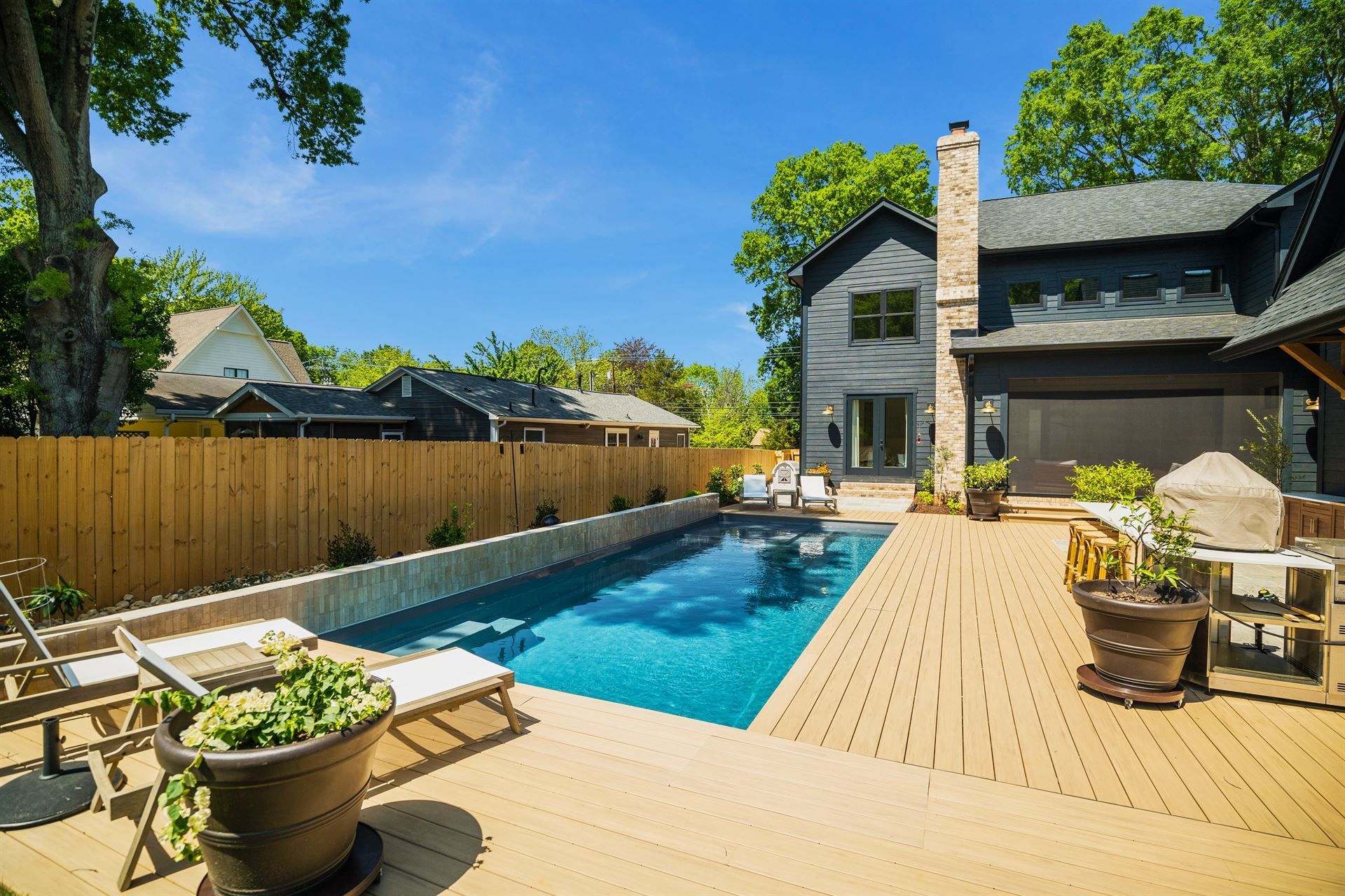 Backyard pool with wooden deck beside a modern house, fenced yard, and potted plants under a clear blue sky