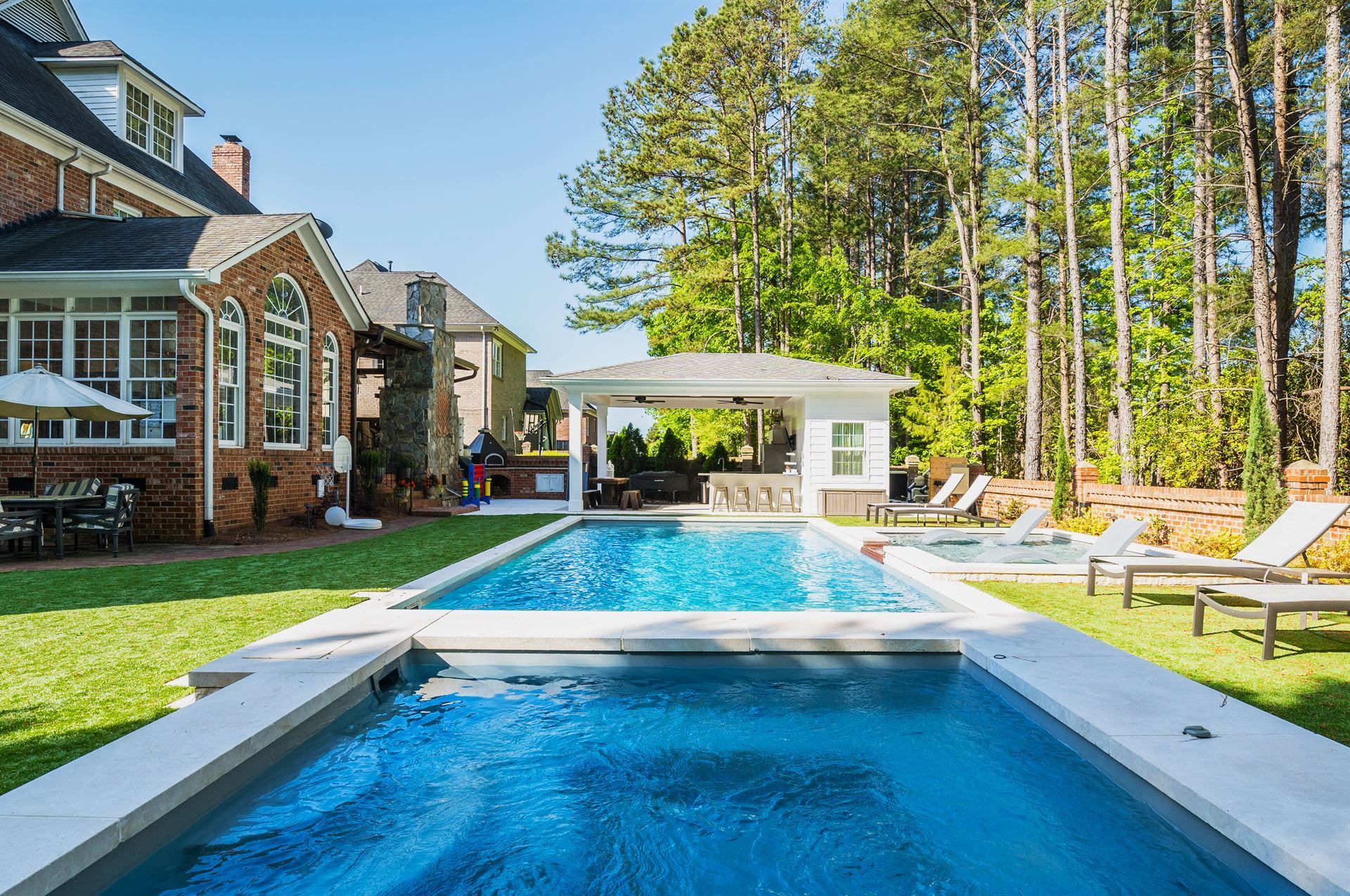 Backyard with long blue pool beside a brick house, lounge chairs, pergola, and tall trees
